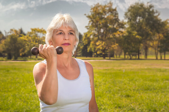 Elderly woman lifts dumbbells while doing fitness in a city park against the backdrop of mountains on a sunny day. Close-up