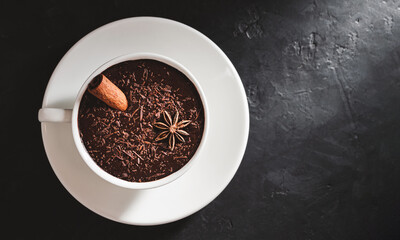 Hot chocolate drink in a white cup with cinnamon stick and star anise on textured dark background, top view with copy-space