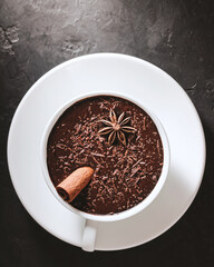 Hot chocolate drink in a white cup with cinnamon stick and star anise on textured dark background, top view with copy-space