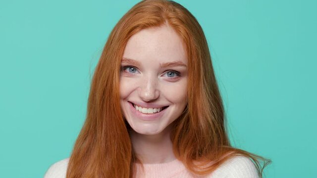 Close up of smiling beautiful ginger redhead young girl wearing white pink sweater posing isolated on blue turquoise color background in studio. People emotions lifestyle concept. Looking camera aside