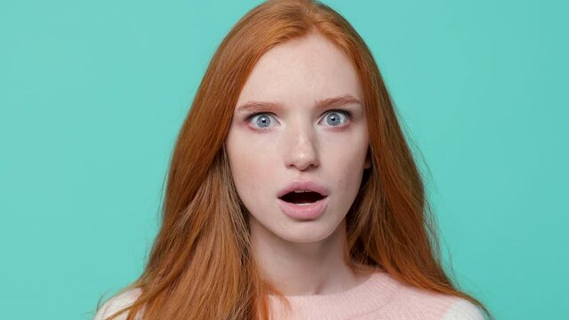 Close up of shocked amazed ginger redhead young girl wearing white pink sweater posing isolated on blue turquoise color background studio. People lifestyle concept. Looking camera keeping mouth open