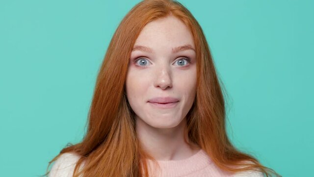 Close up of excited redhead young girl in white pink sweater isolated on blue turquoise color background studio. People lifestyle concept. Looking camera covering mouth with hands showing thumb up