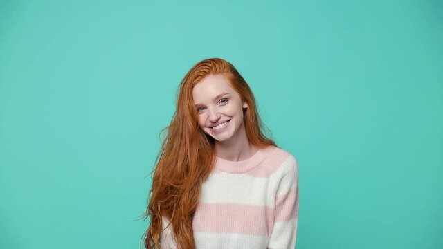 Smiling cheerful ginger redhead young girl wearing white pink sweater posing isolated on blue turquoise color background in studio. People lifestyle concept. Dancing shaking head with fluttering hair