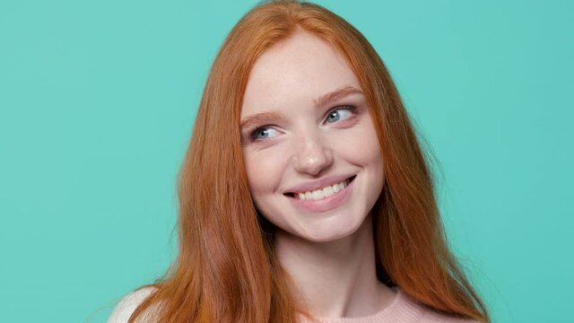 Close up of smiling pensive cute ginger redhead young girl wearing white pink sweater posing isolated on blue turquoise color background in studio. People emotions lifestyle concept. Looking aside up