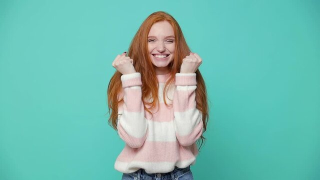 Happy joyful ginger redhead young girl wearing white pink sweater posing isolated on blue turquoise color background in studio. People lifestyle concept. Doing winner gesture clenching fists dancing