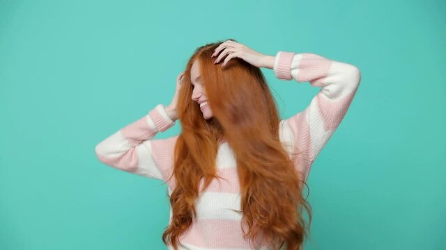 Beautiful cheerful ginger redhead young girl wearing white pink sweater posing isolated on blue turquoise color background in studio. People lifestyle concept. Dancing shaking head with flowing hair