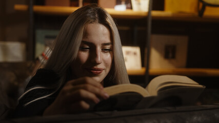 Young blond beautiful woman lying on sofa happily and curiously reading a book or novel