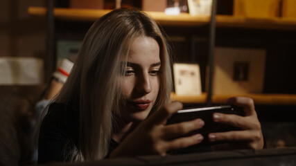 Young blond beautiful woman stretching on the sofa is playing games with smartphone and rejoicing