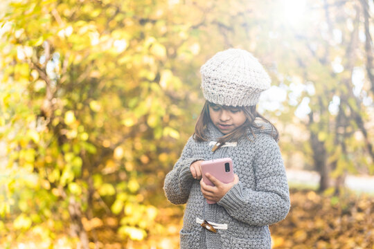 Cute little girl holding smartphone at park during foliage time - Little girl using a phone typing and taking photos of yellow autumnal landscape - Childhood and tech concept in the nature, Copy space