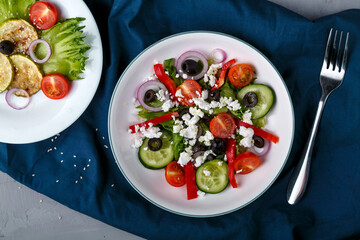 White plate with grilled zucchini with cherry tomatoes on lettuce leaves and Greek salad on a blue napkin with a fork.