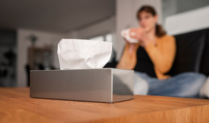 Focus of a stainless steal dispenser with paper handkerchiefs and a young woman in die background holding a mug with tea