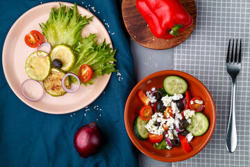 a plate with grilled zucchini on lettuce leaves and a greek salad on a gray background on a blue napkin with a fork near peppers and onions