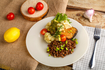 Baked fish with broccoli and lentils in a white plate with rosemary and lemon on a checkered napkin next to a fork and vegetables.
