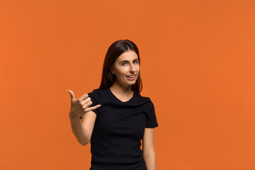 Enjoy distant communication. Positive talkative caucasian woman in black t-shirt makes telephone gesture, smiles pleasantly. Isolated over an orange background