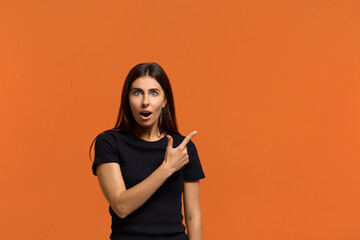 Surprised caucasian woman in black t-shirt with stupefied expression, points with index finger at upper right corner, shows incredible thing, opens mouth. Isolated over an orange background