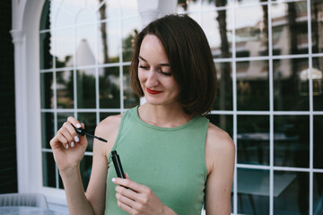 Girl in green top paints eyelashes and holding mascara in her hands on the street. Luxury decorative cosmetics. 