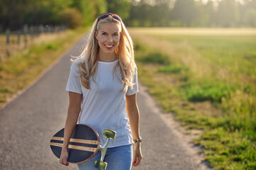 Portrait of a fit beautiful middle-aged woman with an active lifestyle smiling and looking at camera while holding a longboard on a sunny road in the park in summer