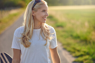 Fit healthy young blond woman carrying a skateboard along a narrow country road looking off the side with a happy smile backlit by the sun