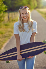 Portrait of a fit beautiful middle-aged woman with an active lifestyle smiling and looking at camera while holding a longboard on a sunny road in the park in summer