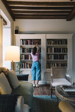 Girl looking for a book in bookcase in a rustic living room at evening.