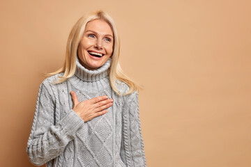 Sincere positive middle aged woman laughs out happily keeps hand on chest smiles broadly has...
