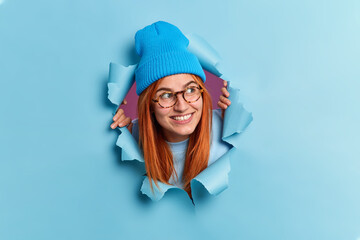 Positive redhead young woman looks away with pleasant smile has curious expression wears hat and...