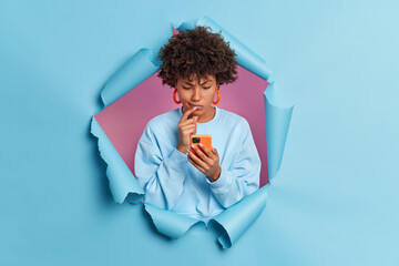 Puzzled dark skinned young Afro American woman looks doubtful concentrated in smartphone display reads comments under post on website wears casual jumper stands in studio through torn paper hole - obrazy, fototapety, plakaty