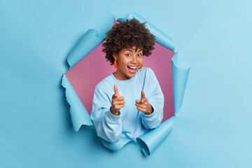 Joyful smiling dark skinned Afro American woman points index fingers at camera makes gun gesture picks you selects someone dressed in casual blue jumper stands through paper torn hole. Join me