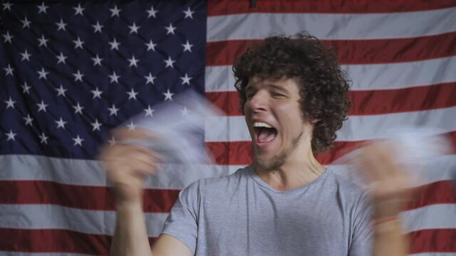 Young guy in background of flag of America throws paper bills with image of American dollars. He's having fun. Model with meek curly hair.High quality 4k footage