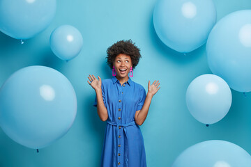 Positive carefree African American woman has festive mood raises hands and looks joyfully up ready for celebration dressed in festive clothing poses against blue background with big balloons © WHstudio Leushin N