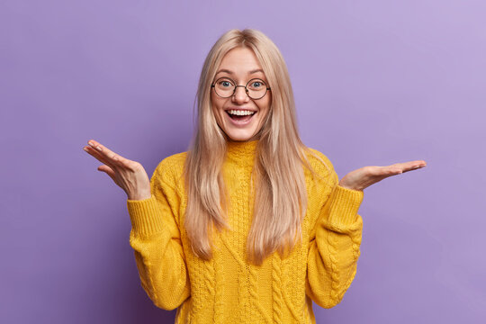 Confused cheerful blonde young woman spreads palms and looks unaware at camera has happy expression wears round spectacles yellow jumper isolated over purple background. Human perception concept