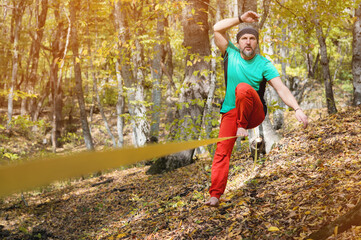 A bearded man in age balances while sitting on a taut slackline in the autumn forest. Outdoor Leisure