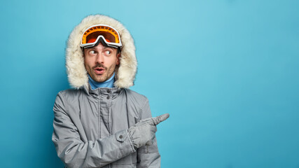 Surprised adult man wears winter jacket and ski goggles has wondered face expression indicates on blank space shows copy space over blue background. Male snowboarder advertises something. Winter time
