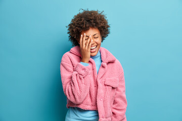 Fototapeta na wymiar Pretty dark skinned woman makes face palm and laughs out joyfully hears good joke dressed in fur pink coat isolated over blue background. Overjoyed Afro American female wears fashionable outerwear
