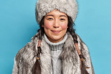 Photo of satisfied young siberia woman with two pigtails rosy cheeks smiles pleasantly at camera dresses for cold polar weather conditions isolated over blue background. Winter time concept. - obrazy, fototapety, plakaty