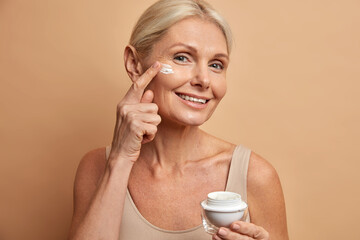 Smiling tender woman holds jar of cream with anti aging effect applies on face has healthy glowing...
