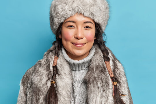 Photo of satisfied young siberia woman with two pigtails rosy cheeks smiles pleasantly at camera dresses for cold polar weather conditions isolated over blue background. Winter time concept.