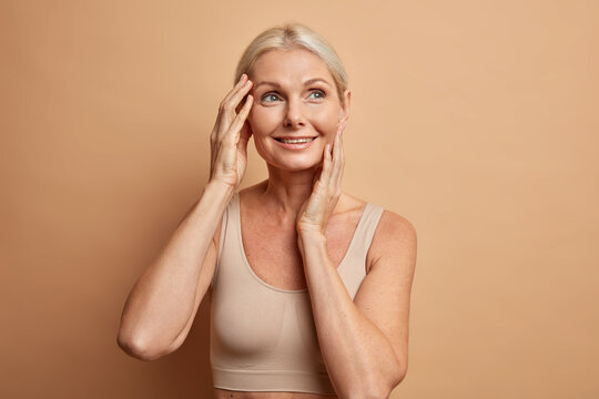 Portrait of good looking middle aged woman with blonde hair touches face and looks thoughtfully away dressed in casual top isolated over brown background. Aging process and beauty time concept