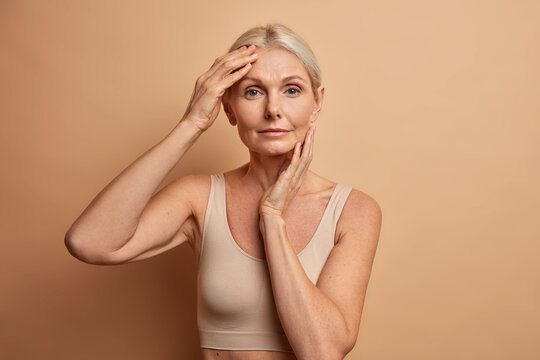 Beautiful middle aged woman touches face has perfect skin after plastic surgery or collagen injections wears minimal makeup wears cropped top isolated over brown studio background. Aging concept