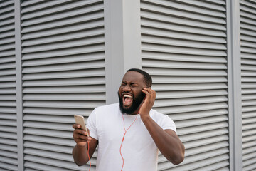 Emotional African American man listening to music outdoors, singing song. Happy hipster guy wearing...