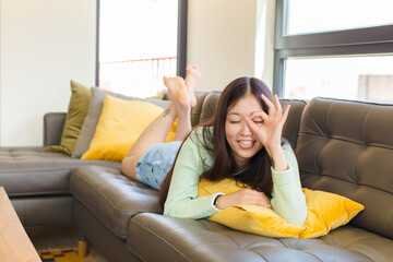 young asian woman smiling happily with funny face, joking and looking through peephole, spying on...