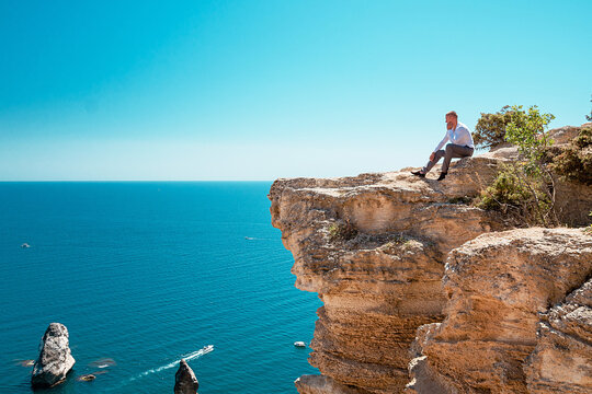 A pensive young man sits at a cliff. Portrait of a guy by the sea.