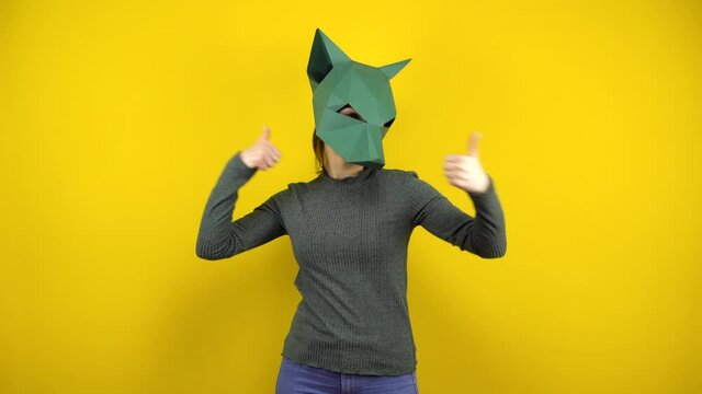 A young woman in a cardboard jackal mask shows her thumbs up on a yellow background. Woman in green sweater and mask.