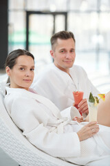 Young restful couple in white bathrobes having tropical cocktails in spa center