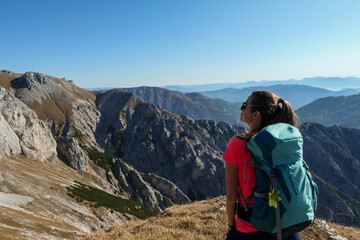 Fototapeta premium A woman with big blue backpack enjoying the view on valley from top of a mountain in Hochschwab region in Austrian Alps. The flora overgrowing the slopes is turning golden. Freedom. She is happy