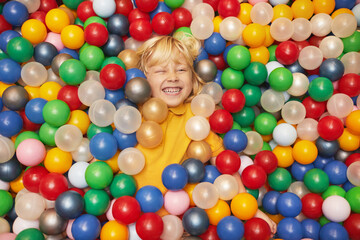 Happy little boy lying in pool with colored balls and enjoying the game