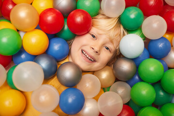 Fototapeta na wymiar Close-up of happy boy with blond hair smiling at camera while lying among colored balls and playing
