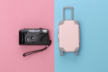 Travel concept. Mini plastic travel suitcase and camera on pink blue pastel background. Minimal style. Top view, flat lay