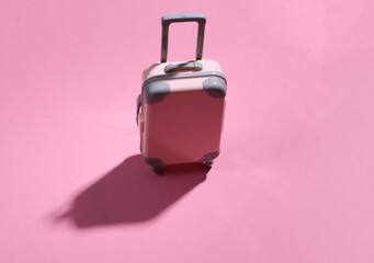 Travel minimalism. Mini plastic travel suitcase on pink background with deep shadow. Minimal style