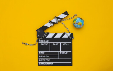 Movie clapperboard wrapped in steel chain and globe on yellow background. Ban on filmmaking. Top...